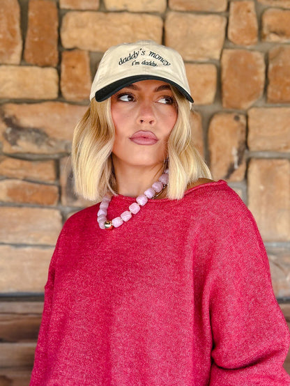 A woman with blonde bob-cut hair wearing a cream baseball cap with black embroidery that reads 'Daddy's money is mine', a bright pink knit sweater, and a pink beaded necklace, standing in front of a rustic stone wall.
