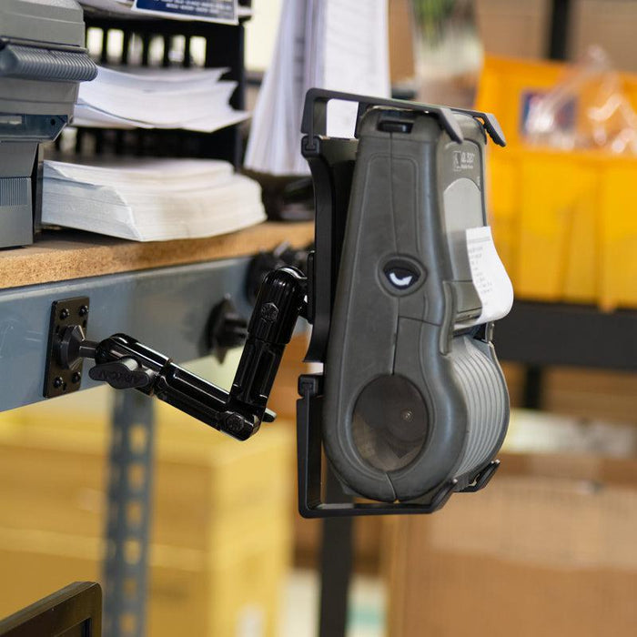 A Portable Mobile Printer Car Truck Drill Base Mount for Zebra, Epson, and Brother Printers is secured to the edge of a metal workbench, holding a handheld label printer; paperwork and office supplies are in the background.