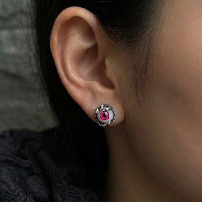 A close-up of a person's ear wearing a gothic sterling silver stud earring featuring a realistic red evil eye design held in a textured silver claw setting.
