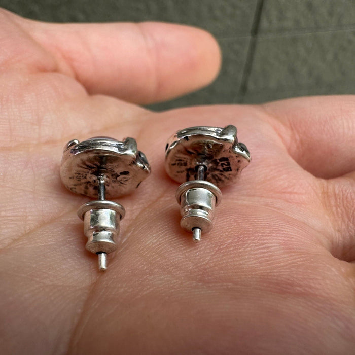 Reverse side of two sterling silver stud earrings in a palm, showing the textured metal base, posts, and butterfly backs.