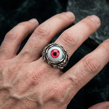 A silver ring featuring a red eyeball design worn on a hand against a dark background.