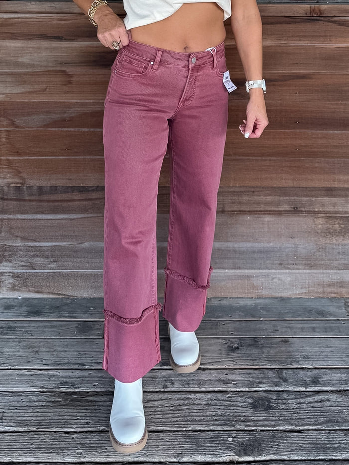 A person wearing dusty rose wide-leg cropped jeans with frayed hem details and rolled cuffs, paired with white ankle boots, standing against a rustic wooden plank wall.