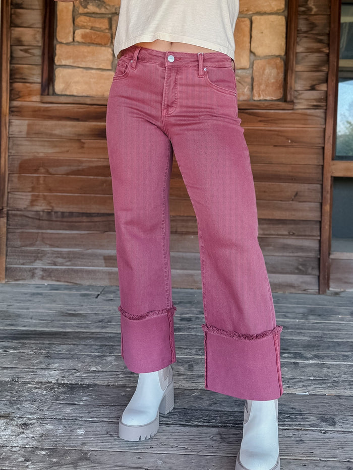A person wearing dusty rose pink wide-leg cropped jeans with rolled cuffs, paired with white platform boots, standing on a weathered wooden deck in front of a rustic stone and wood wall.