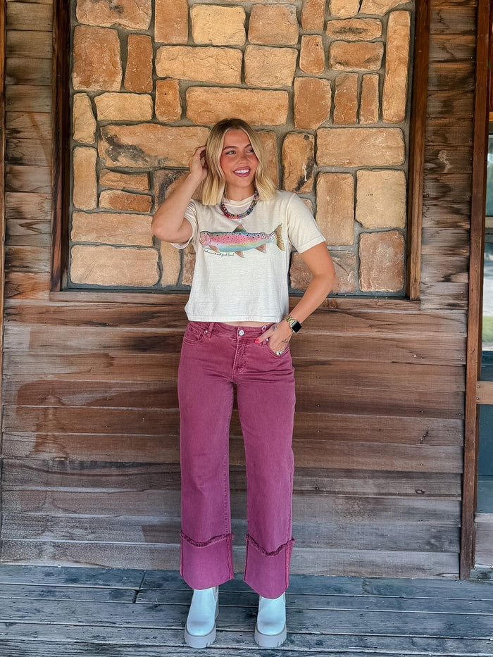 A woman stands on a wooden porch in front of a stone wall, wearing a cream graphic t-shirt with a fish design and dusty rose corduroy pants.