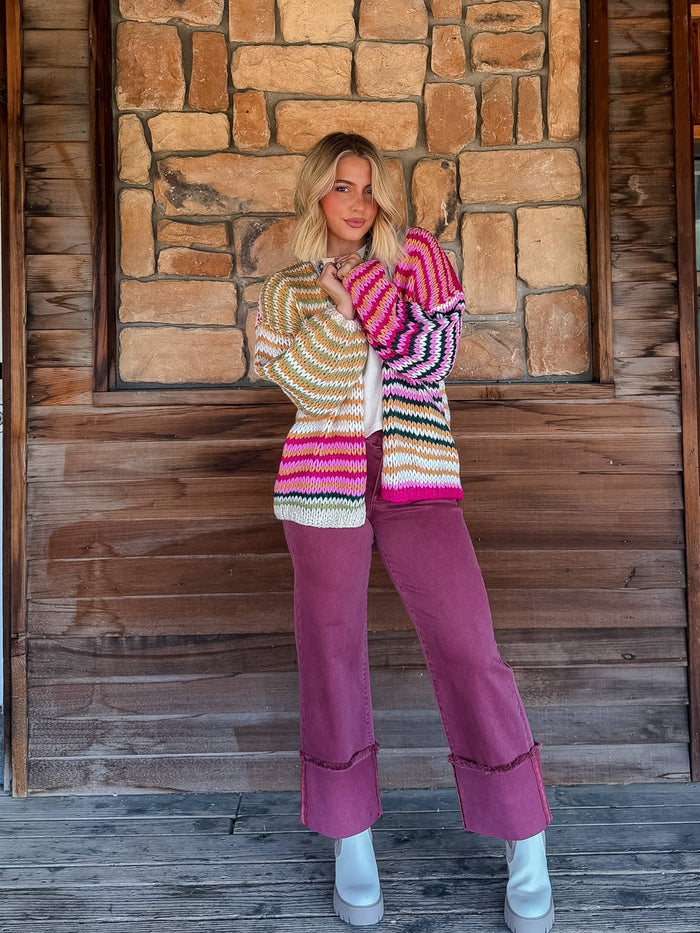 A woman stands on a rustic wooden porch in front of a stone wall, wearing a colorful striped knit cardigan and mauve wide-leg pants with white boots.