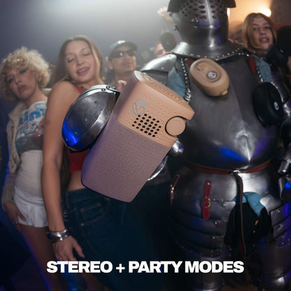 A portable Session 900 speaker with a textured taupe body is shown in a crowded party scene, with people and a knight in armor in the background. The text 'STEREO + PARTY MODES' is displayed at the bottom. | Primer