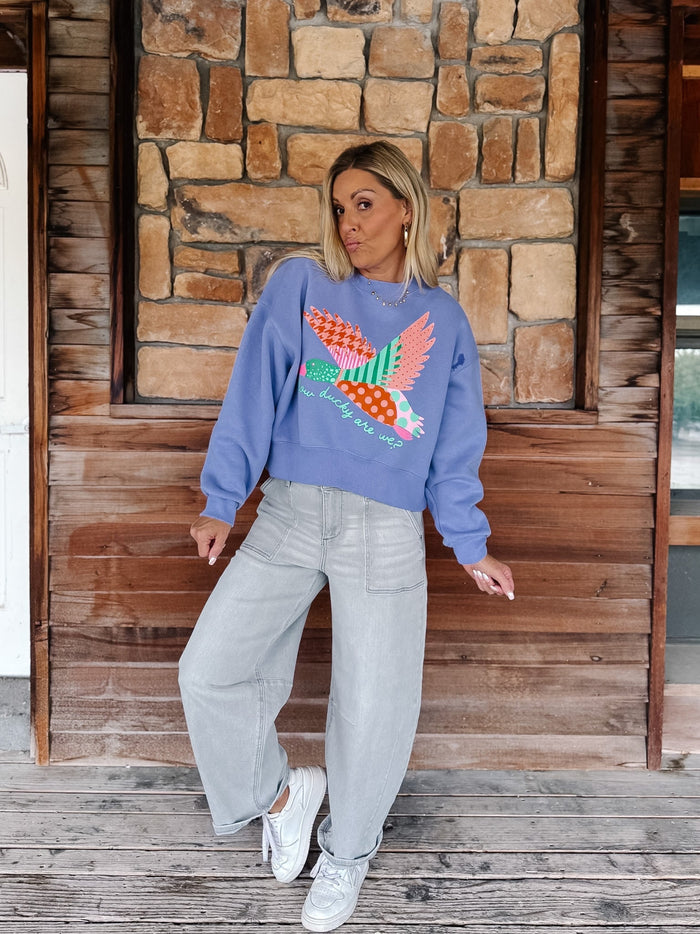 A woman stands on a wooden porch wearing a lavender crewneck sweatshirt with a colorful embroidered bird design, paired with light gray wide-leg jeans and white sneakers.