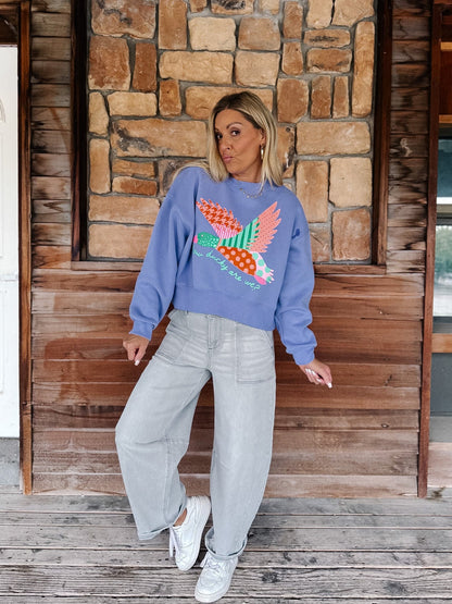 A woman stands on a wooden porch wearing a lavender crewneck sweatshirt with a colorful embroidered bird design, paired with light gray wide-leg jeans and white sneakers.