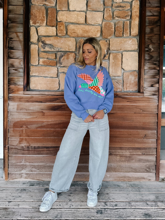 A woman stands on a wooden porch in front of a stone wall, wearing a blue crewneck sweatshirt with a colorful embroidered bird graphic, light-wash wide-leg jeans, and white sneakers.