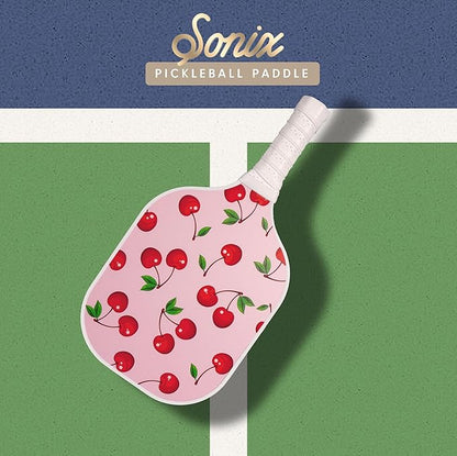 Top-down view of Sonix pickleball paddle with pink cherry print and white wrapped handle on a green court