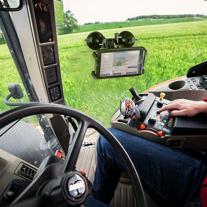 A person operates a modern tractor using control panels and a touchscreen, green fields visible outside, while a Slim-Grip® Double Suction Windshield Tablet Mount secures an extra screen to the dashboard.