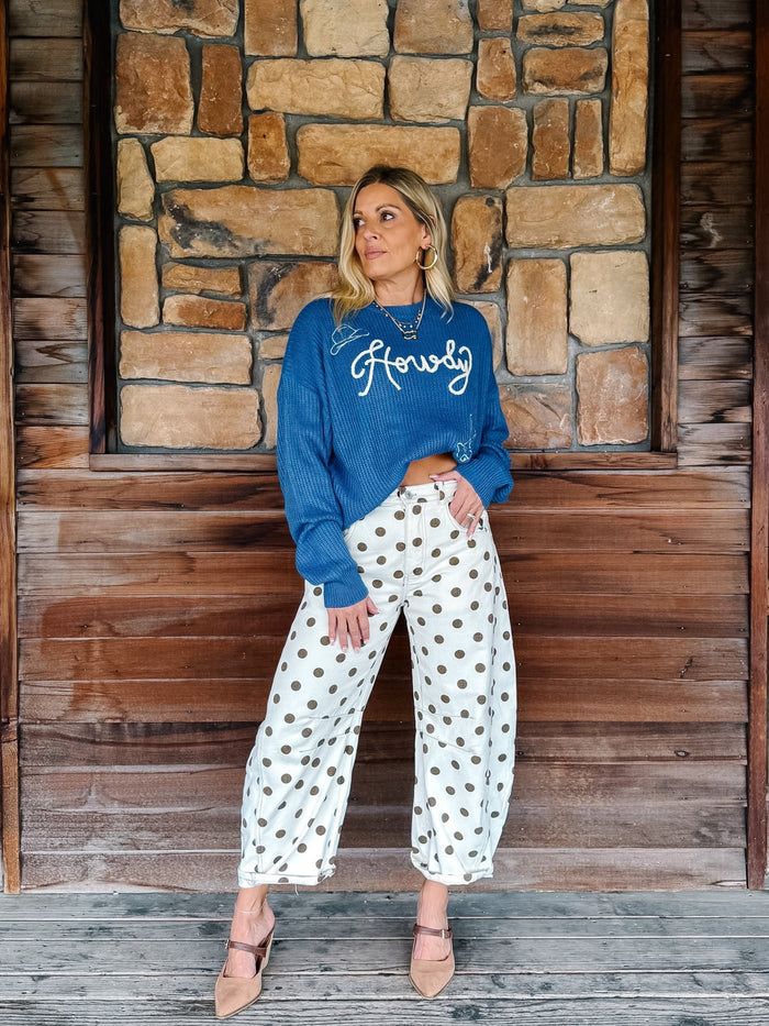 A woman stands against a rustic stone and wood wall wearing a blue crewneck sweatshirt with 'Honey' embroidered in white script, white wide-leg pants with brown polka dots, and tan pointed-toe flats.