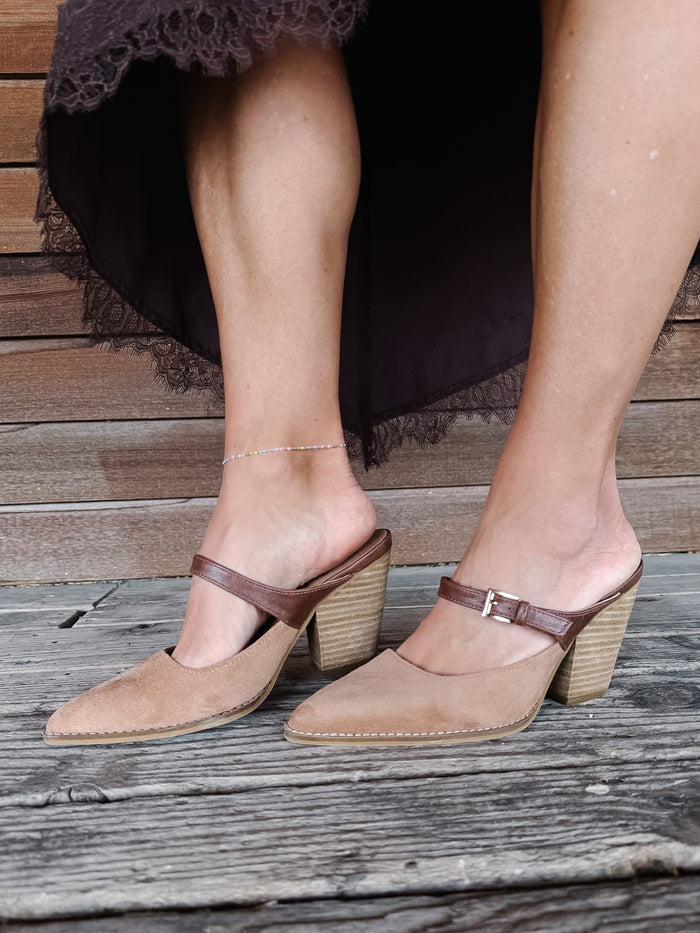 A pair of women's pointed-toe mule heels in tan suede with brown straps and stacked wood-look block heels, shown on a model standing on weathered wooden planks.