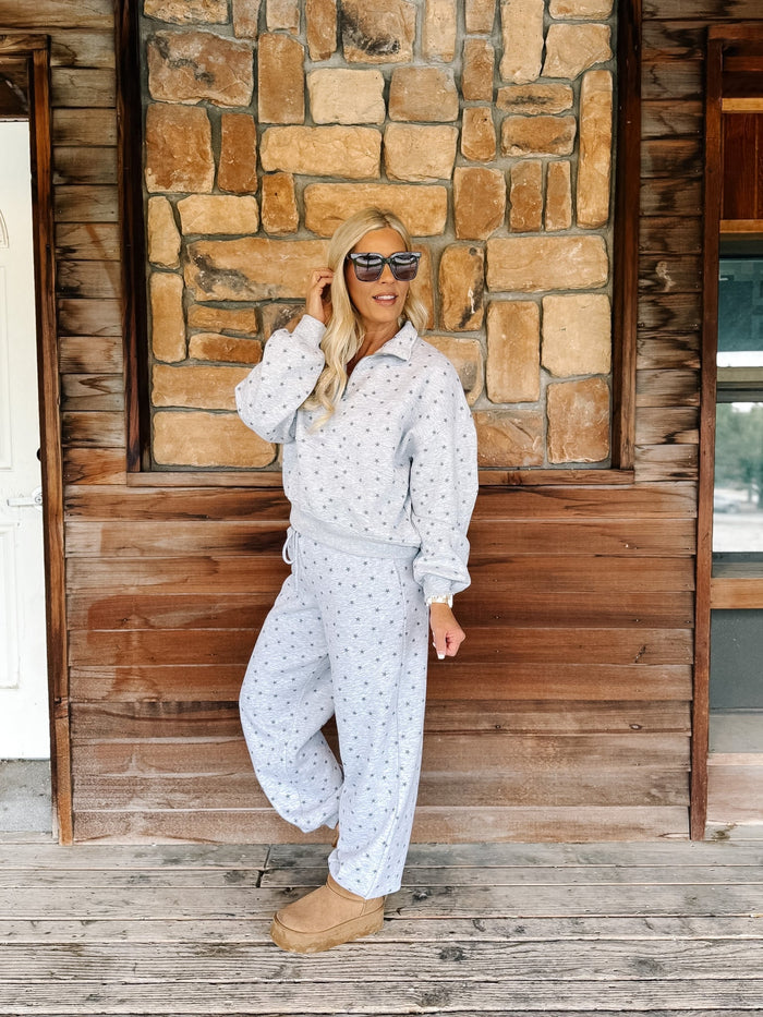 A person wearing a light gray and white star patterned sweatshirt and matching sweatpants, paired with tan suede boots, standing on a rustic wooden porch in front of a stone wall.