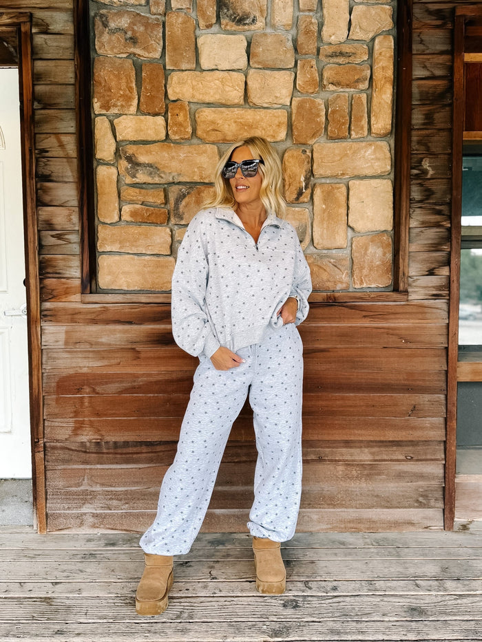 A person wearing a light gray and white star patterned sweatshirt and matching sweatpants, paired with tan suede boots, standing on a rustic wooden porch in front of a stone wall.