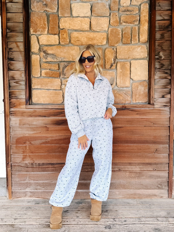 A person wearing a light gray and white star patterned sweatshirt and matching sweatpants, paired with tan suede boots, standing on a rustic wooden porch in front of a stone wall.