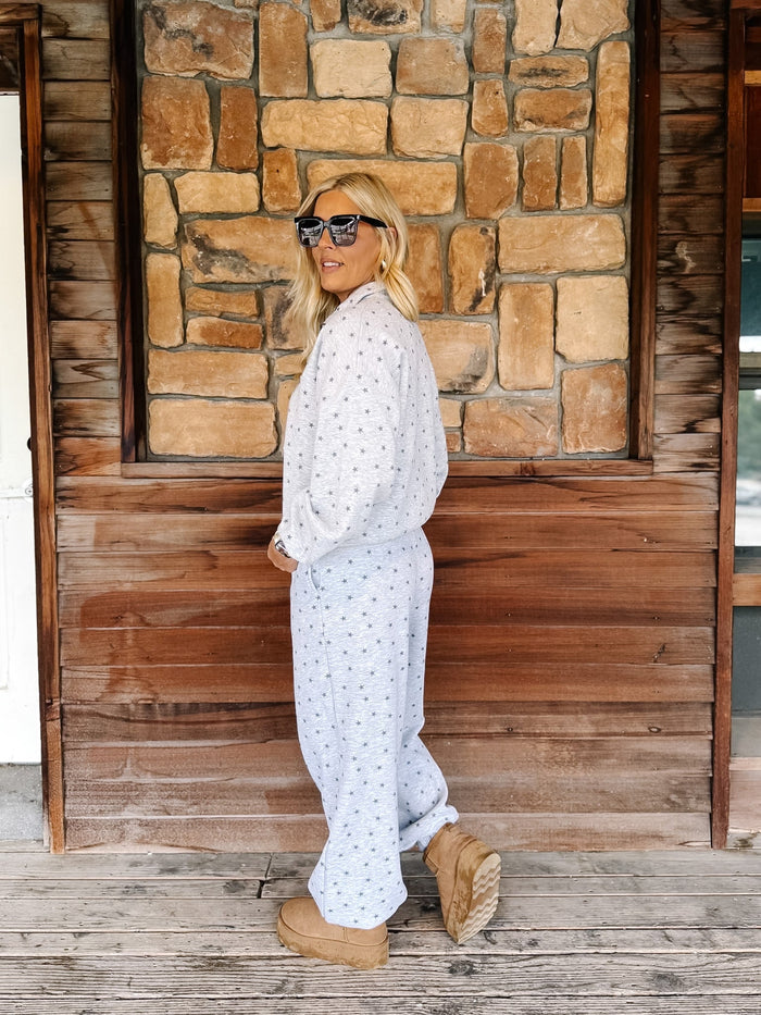 A person wearing a light gray and white star patterned sweatshirt and matching sweatpants, paired with tan suede boots, standing on a rustic wooden porch in front of a stone wall.