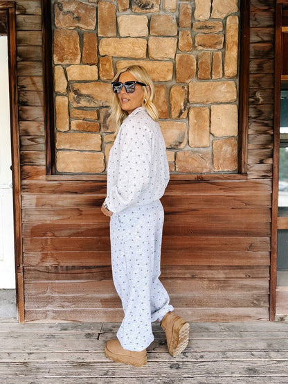 A person wearing a light gray and white star patterned sweatshirt and matching sweatpants, paired with tan suede boots, standing on a rustic wooden porch in front of a stone wall.