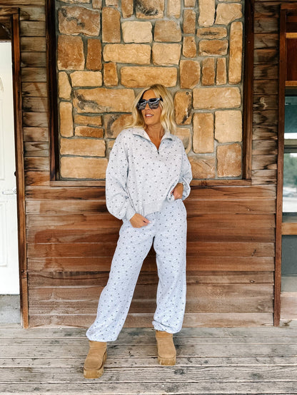 A woman wearing a light heather grey  star patterned matching lounge set stands on a wooden porch in front of a stone wall