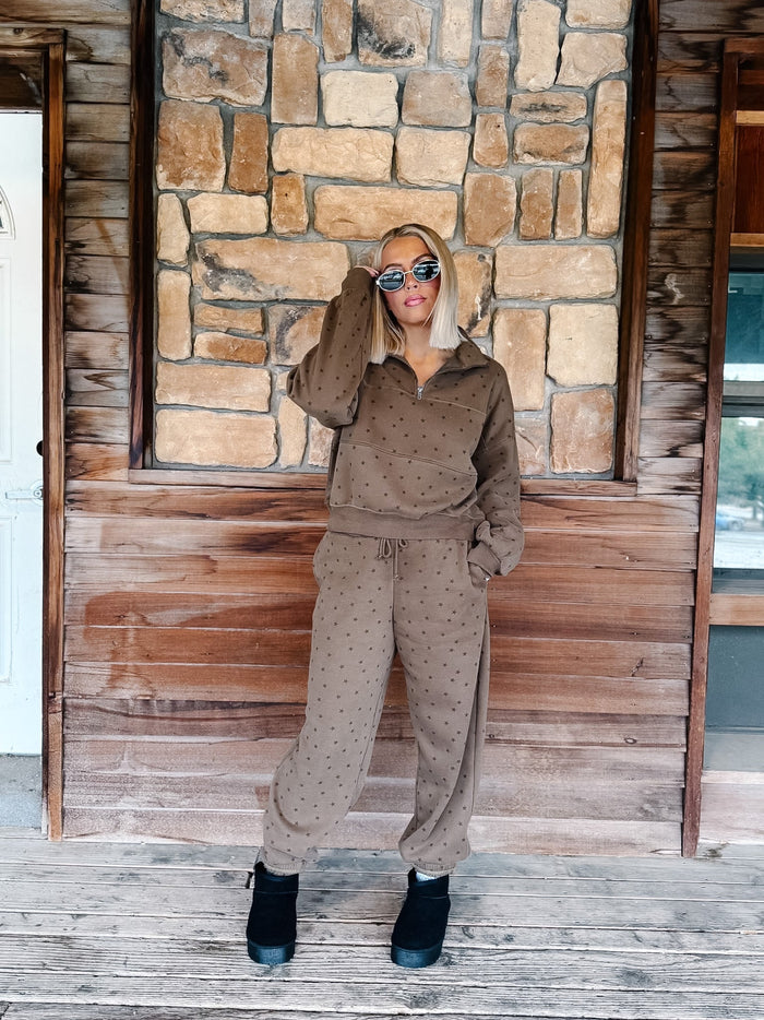A woman with blonde hair and sunglasses wearing an mocha brown star-patterned half-zip sweatshirt and matching sweatpants, standing in front of a stone wall.