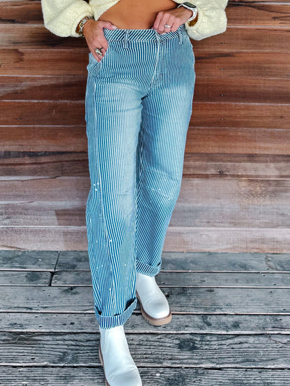 A person wearing light blue striped jeans with white boots, standing on a weathered wooden deck in front of a wooden wall.