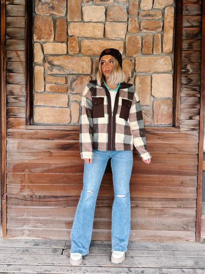 A woman stands in front of a rustic stone and wood wall wearing a brown, beige, and white plaid zip-up hoodie with a black beanie, light blue distressed jeans, and white sneakers.
