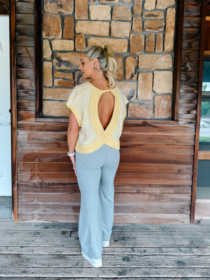 A woman wearing a yellow and heather oatmeal striped short-sleeve  top with gray drawstring sweatpants, posing in front of a rustic stone wall.