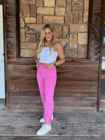 A woman stands on a wooden deck in front of a rustic stone and wood wall, wearing a light blue sleeveless top, bright pink skinny jeans, and white sneakers.