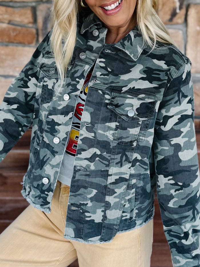 A woman wearing a cropped camouflage denim jacket with a button-front closure and chest flap pockets, paired with beige pants, shown against a brick wall background.