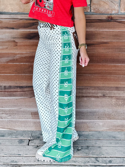 A person wearing wide-leg white pants with black polka dots and a green football-field stripe down the side, paired with a red graphic t-shirt and white sneakers, standing against a rustic wooden wall.