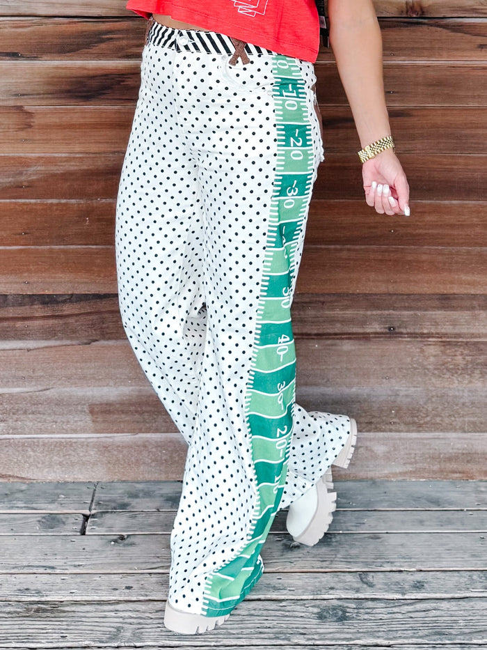 White wide-leg pants with a black polka dot pattern and a green measuring tape graphic running down the side, modeled against a rustic wooden plank background.