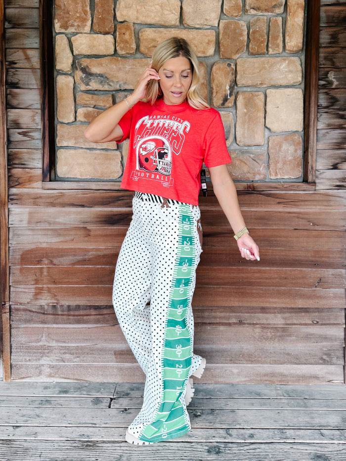 A woman stands on a rustic wooden porch in front of a stone wall, wearing a red graphic t-shirt and white wide-leg pants with green and white polka dots and a green side stripe.