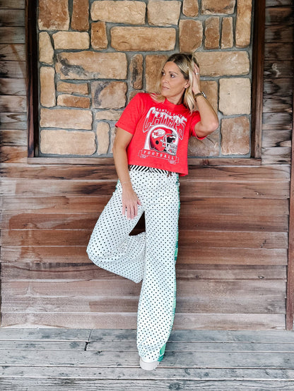 A woman posing in a red graphic t-shirt and white polka dot pants against a rustic wooden and stone wall background.