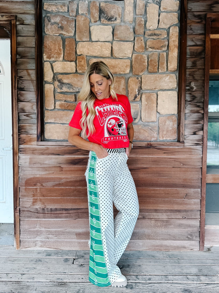 A woman stands on a rustic wooden porch in front of a stone wall, wearing a red graphic t-shirt and white wide-leg pants with green and black polka dots.