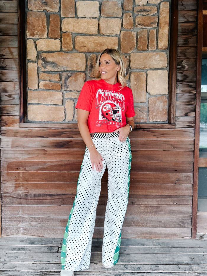 A woman stands on a wooden porch in front of a stone wall, wearing a red graphic t-shirt and white wide-leg pants with black polka dots and green side accents.