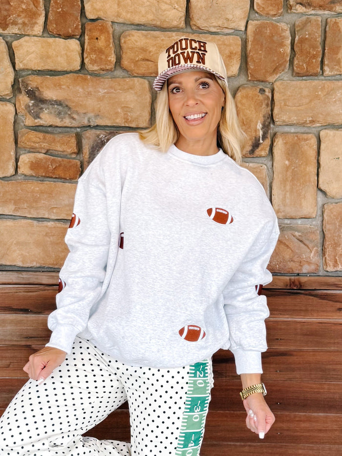 A woman wearing a white heather crewneck sweatshirt with football graphics, white polka dot pants, and a beige cap that reads 'MUCH DOM', posing in front of a rustic stone wall.