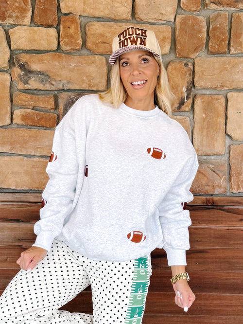 A woman wearing a white heather crewneck sweatshirt with football graphics, white polka dot pants, and a beige cap that reads 'MUCH DOM', posing in front of a rustic stone wall.