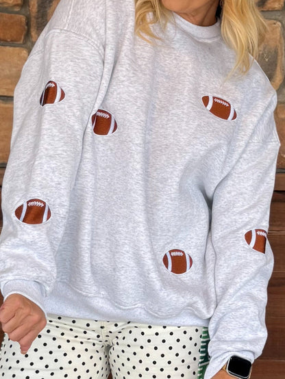 A person wearing a light heather gray sweatshirt with brown and white football patches sewn onto the front, paired with white pants with black polka dots, standing against a brick wall.