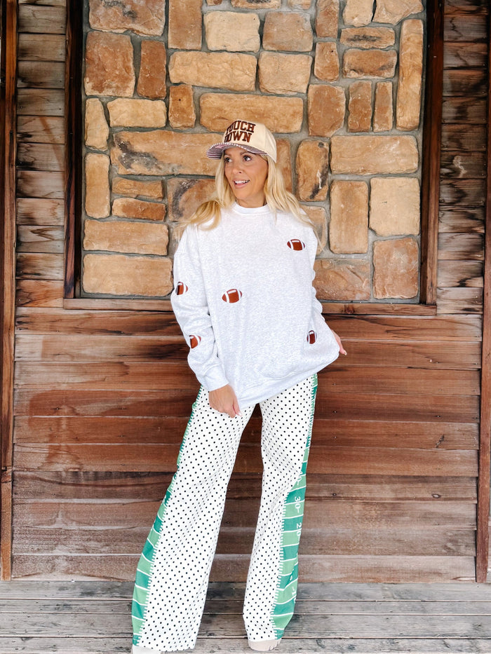 A woman stands in front of a rustic stone and wood wall wearing a white sweatshirt with football patch details, white wide-leg pants with green striped side panels and black polka dots, and a beige baseball cap that reads 'MAKE YOUR OWN'.