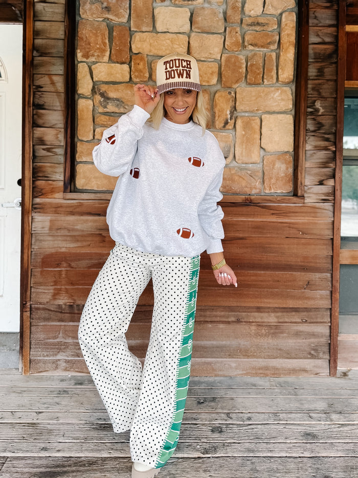 A woman stands on a wooden porch in front of a stone wall, wearing a light gray long-sleeve sweatshirt with football motifs, white wide-leg pants with green side stripes and white polka dots, and a beige cap with 'TOUCH DOWN' text.