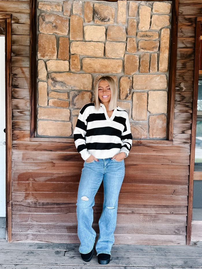A woman stands on a wooden porch in front of a stone wall, wearing a black and white horizontally striped knit sweater and light blue distressed jeans.