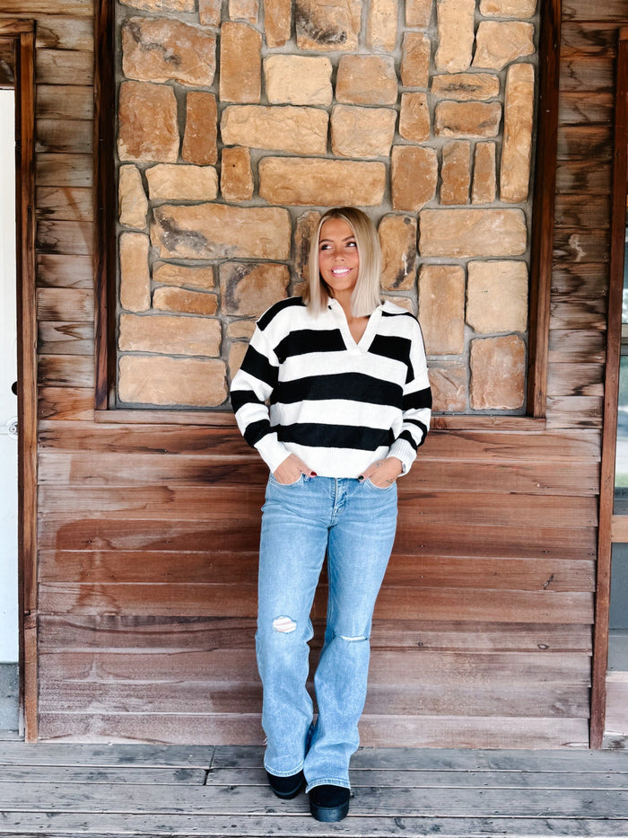 A woman stands on a wooden porch in front of a stone wall, wearing a black and white striped long-sleeve polo sweater, light blue distressed jeans, and black shoes.
