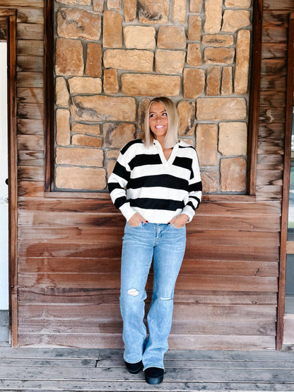 A woman stands on a wooden porch in front of a stone wall, wearing a black and white striped long-sleeve polo sweater, light blue distressed jeans, and black shoes.