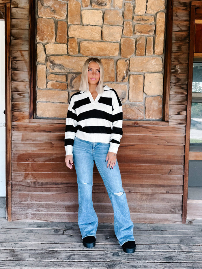 A woman stands on a wooden porch in front of a stone and wood wall, wearing a black and white horizontally striped knit sweater, light blue distressed jeans, and black shoes.