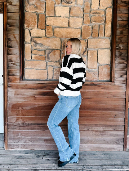 A woman with blonde hair stands on a wooden porch in front of a stone and wood wall, wearing a black and white horizontally striped knit sweater and light blue jeans.