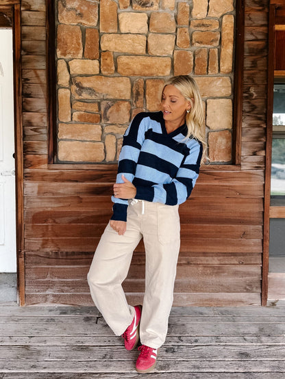A woman stands on a wooden porch in front of a stone wall, wearing a light blue and navy striped long-sleeve knit sweater, cream wide-leg pants, and bright pink sneakers.