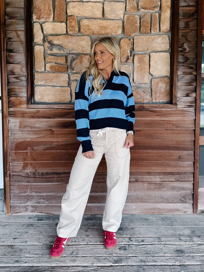 A woman stands on a rustic wooden porch in front of a stone wall, wearing a blue and navy striped long-sleeve knit sweater, cream wide-leg sweatpants, and pink sneakers.