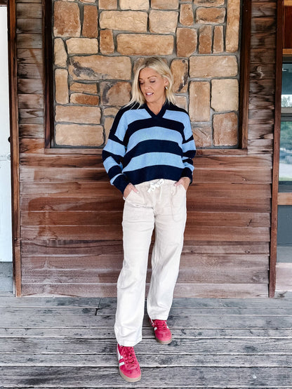 A woman stands on a wooden deck in front of a rustic stone and wood wall, wearing a navy and light blue striped knit sweater, cream drawstring pants, and pink sneakers.