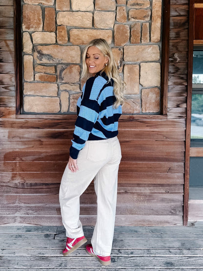 A woman stands on a wooden porch in front of a stone wall, wearing a blue and black striped knit sweater, off-white wide-leg pants, and pink platform sneakers.