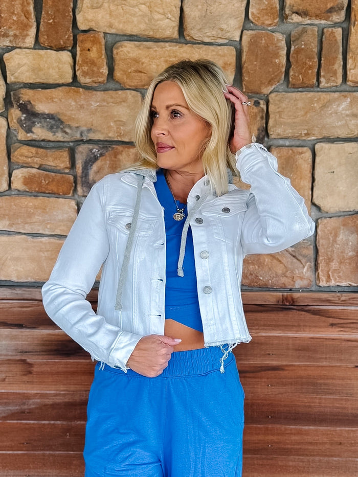 A woman posing in front of a rustic stone wall, wearing a light silver-white cropped denim jacket over a bright blue crop top and matching blue sweatpants.